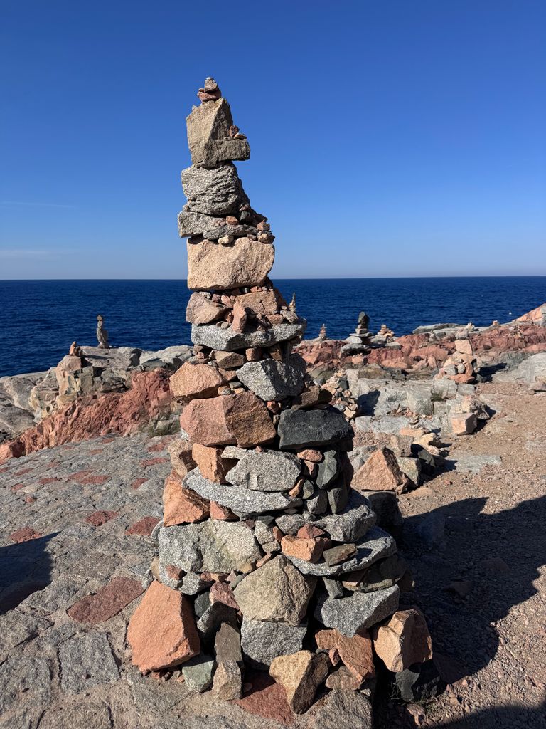 A stunning cairn of stacked stones stands against the coastal backdrop of Sardinia's blue Mediterranean Sea.