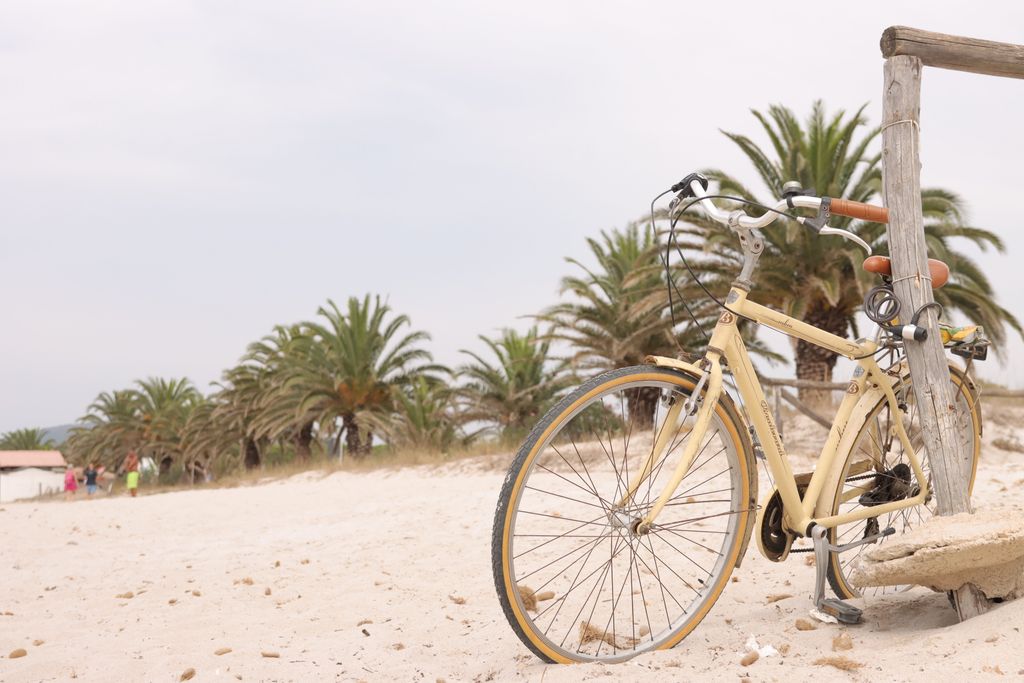 A vintage yellow bicycle leans against a wooden post on a sandy beach with palm trees.