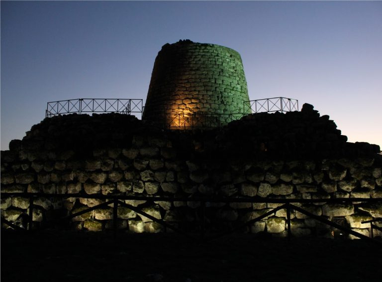 Una fotografia drammatica al tramonto del Nuraghe Santu Antine. La pietra scura contrasta con il cielo infuocato di arancione e viola. In primo piano, erba selvatica mossa dal vento. Atmosfera epica e misteriosa. Alta definizione